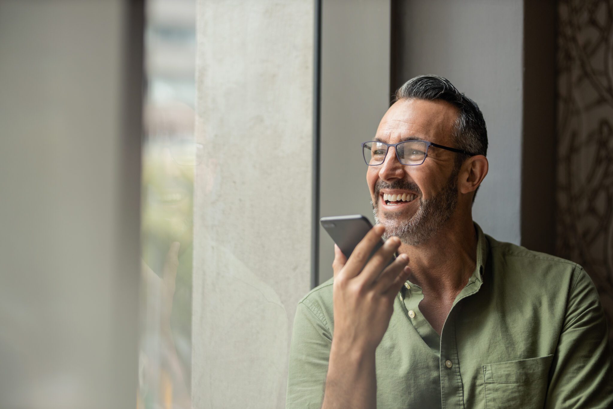 Smiling mature man with beard talking on phone next to sunny window